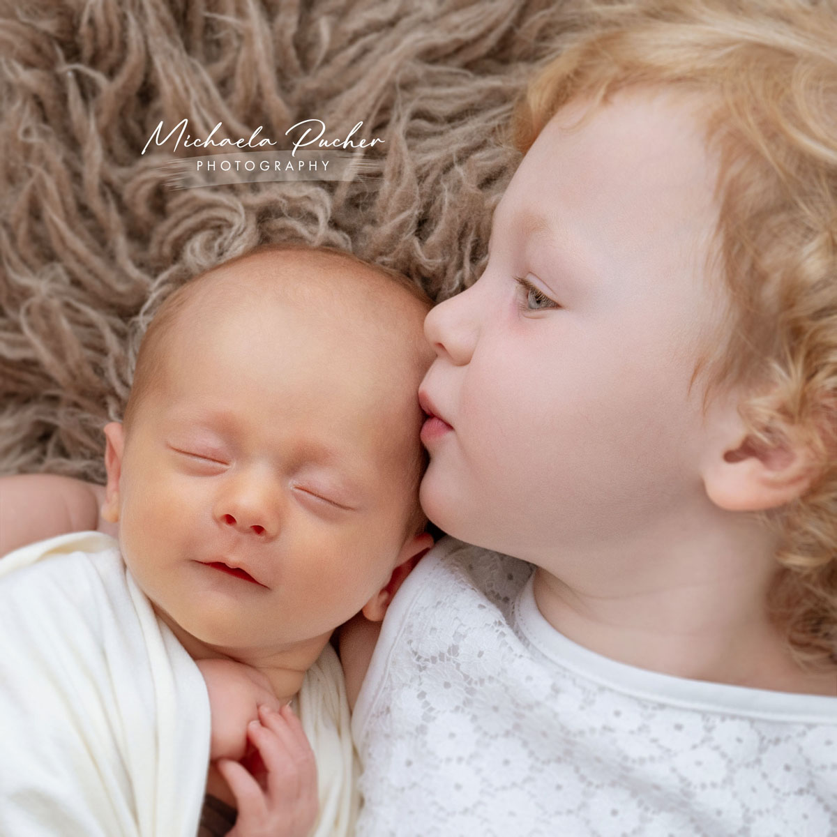 Älteres Geschwisterkind liegt neben dem schlafenden Neugeborenen und gibt ihm einen sanften Kuss auf die Stirn.